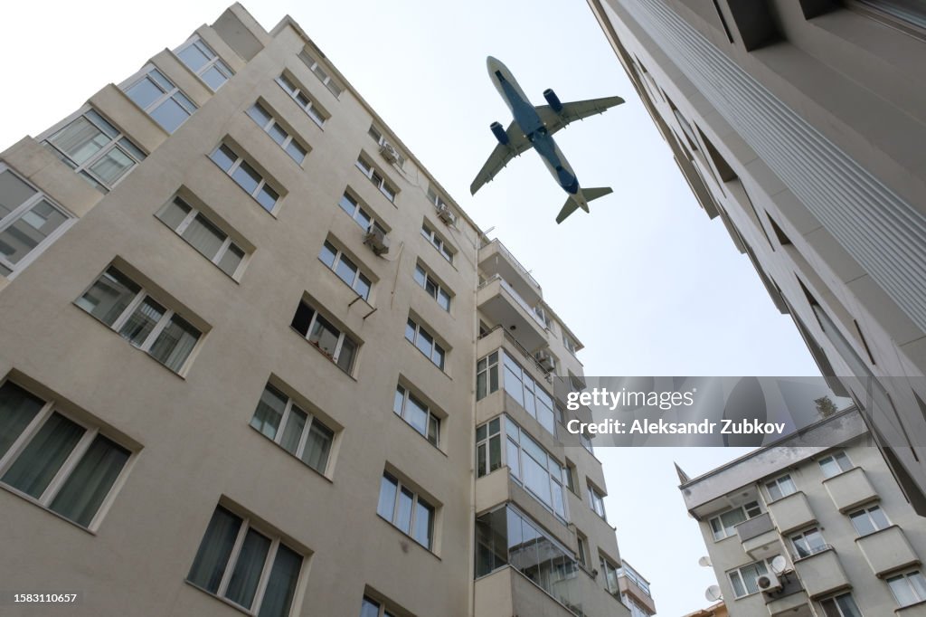 A passenger plane flies low over a house or building in a residential area. Bottom view. The plane is on the background of a blue cloudless sky, landing or taking off near the city. Tourist destinations.