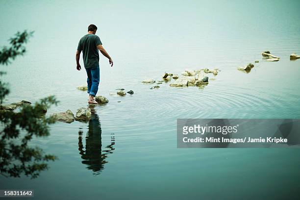 a man walking barefoot across stepping stones away from the shore of a lake. - stepping stones stockfoto's en -beelden