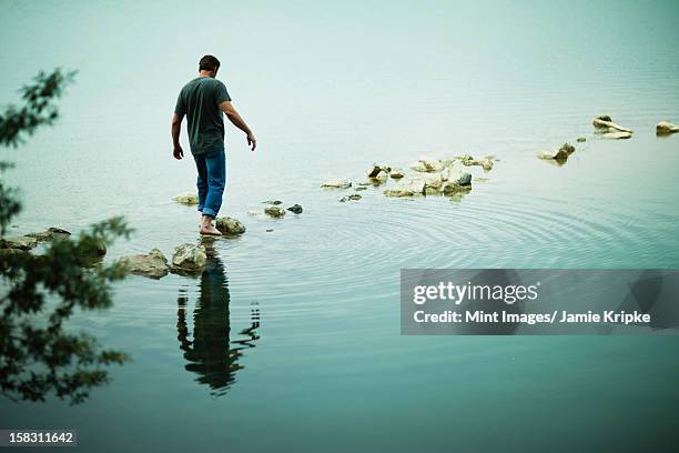 a man walking barefoot across stepping stones away from the shore of a lake. - stepping stone stock pictures, royalty-free photos & images