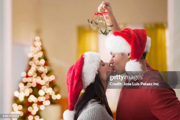 couple in santa hats kissing underneath mistletoe - mistletoe kiss stockfoto's en -beelden