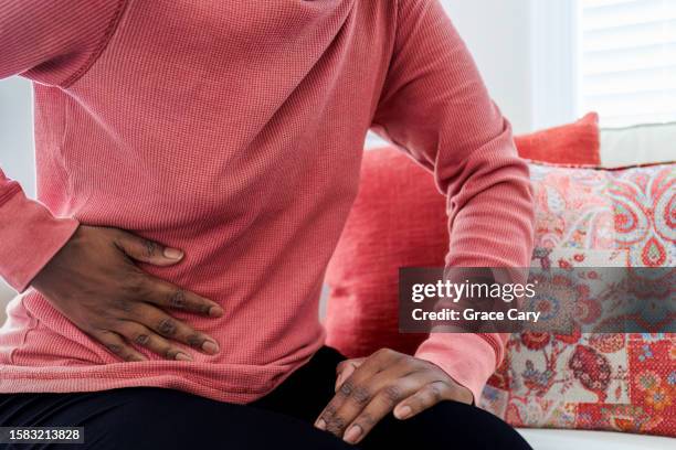 woman sits on couch with abdominal pain - fibroids stockfoto's en -beelden