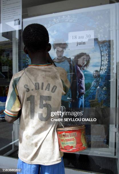 Young beggar wearing a jersey bearing the name of the international Senegalese football player Salif Diao, carrying a metal bin looks at Harry...