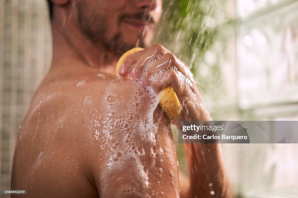 Unrecognizable man taking a shower, using sponge to rub his shoulder.