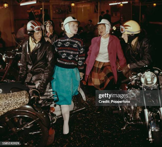 Two couples adorn crash helmets as they stand astride motorcycles outside a biker's cafe, the men wear leather biker jackets, the women wear skirts,...