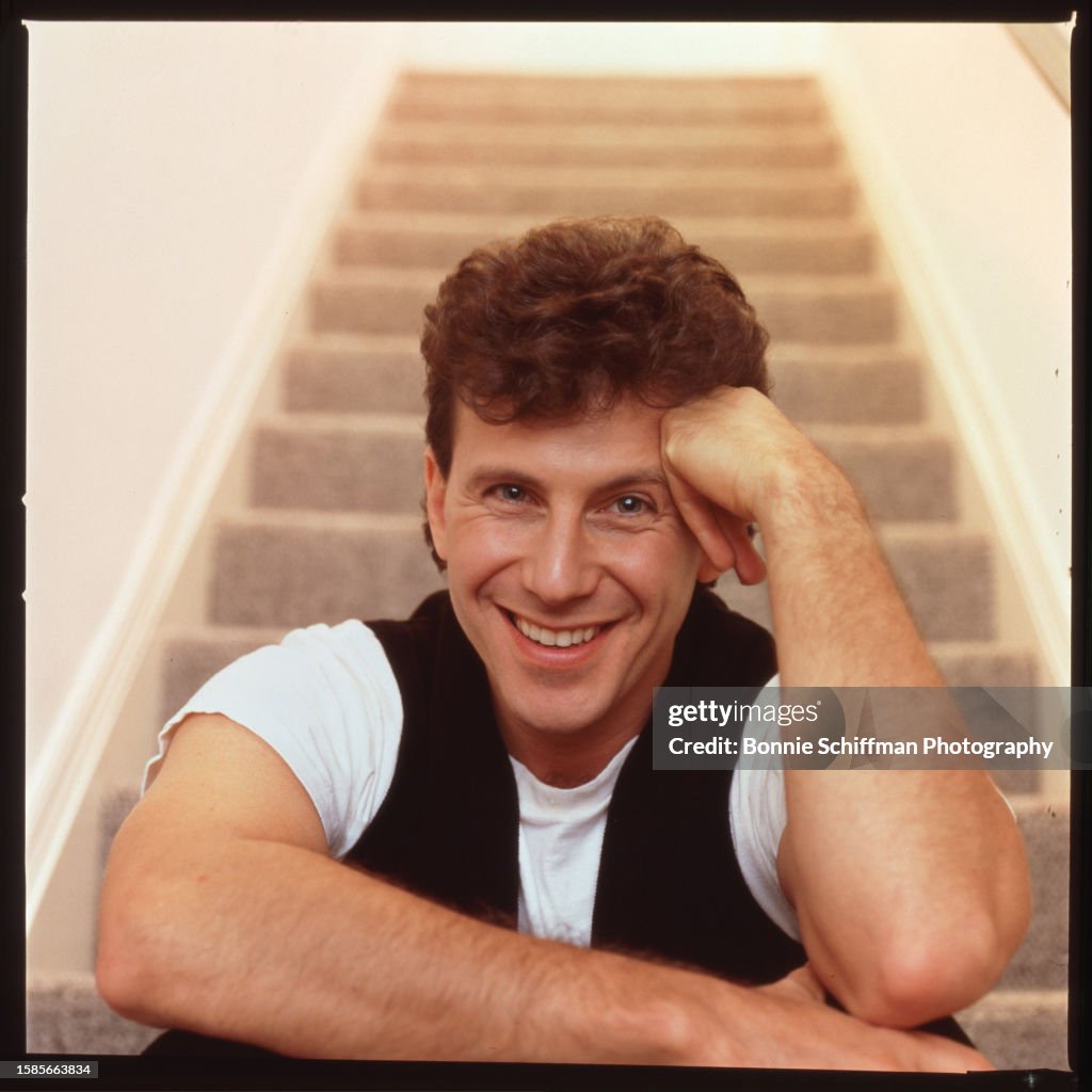 Comedian Paul Reiser sits at bottom of steps and smiles with his hand ...