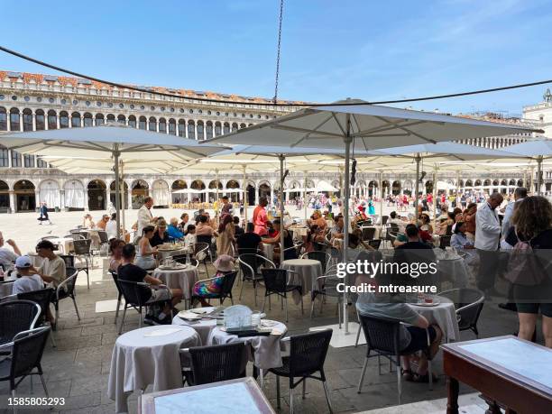 close-up image of crowds of tourists dining al fresco under parasols in piazza san marco (st mark's square) opposite the palazzo ducale (doge's palace), clear blue sunny sky, focus on foreground - waterfront cafe stock pictures, royalty-free photos & images