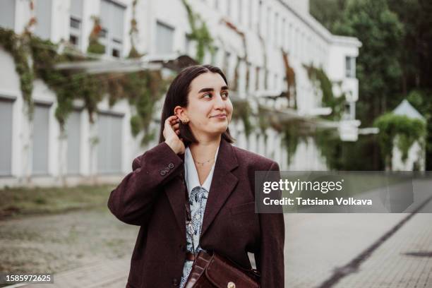 portrait of woman walking across university campus, straightening brown hair and looking away with modest smile. - tímido imagens e fotografias de stock