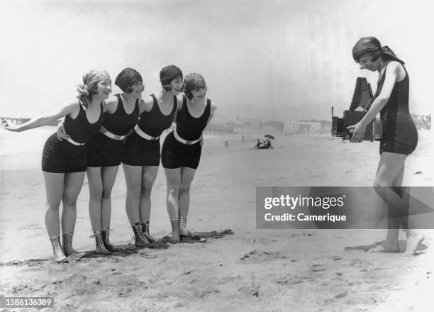 Four of Mack Sennett's 'Bathing Beauties' in one piece costumes being photographed by a fifth on the beach.