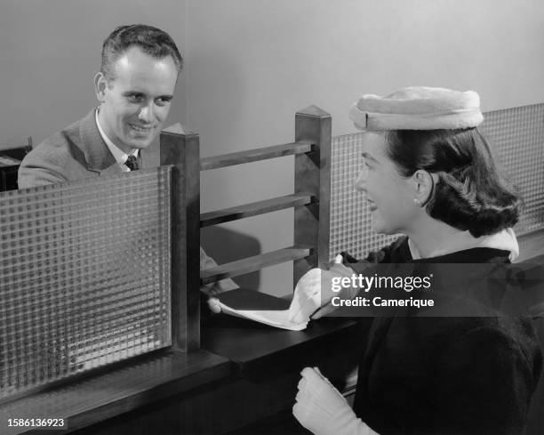 Woman in hat and gloves passes a slip to teh teller in a bank.