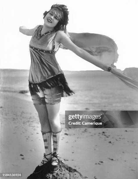 One of the Mack Sennett 'Bathing Beauties' buffeted by the sea breeze as she stands on a mound of sand.