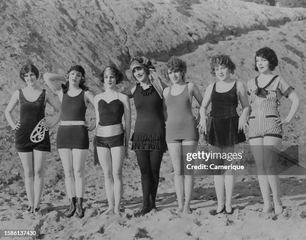 Seven of Mack Sennett's 'Bathing Beauties' in various one piece bathing suits stand posed in a row on a beach.
