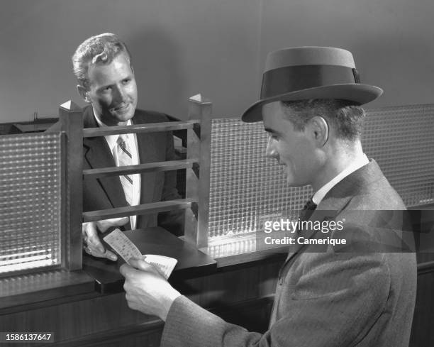 Man in suit and hat looks at a slip that a bank teller has just given him.