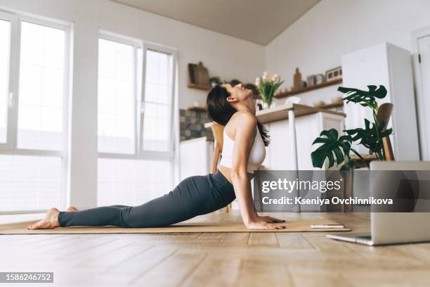 fit woman in sportswear doing yoga on exercise mat at home using laptop watching tutorial. female coach has a virtual yoga class on computer. healthy and active lifestyle concept. online home training - sporting training and press conference stock pictures, royalty-free photos & images