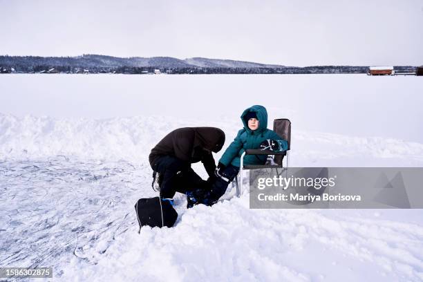 a father puts his son's skates on a makeshift skating rink on the lake. - figure skating child stock pictures, royalty-free photos & images