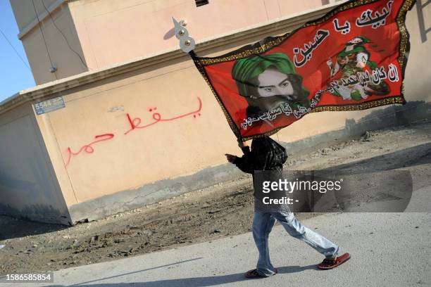 Bahraini Shiite Muslim youth holding a flag with the picture of Imam Hussein, grandson of Islam's Prophet Mohammed, walks past wall graffiti that...