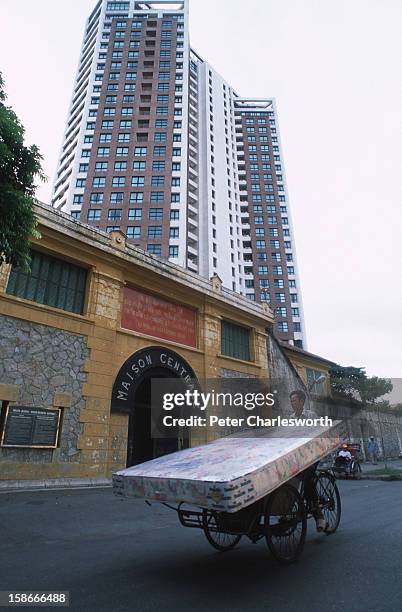 Man with a mattress perched on his cyclo rides past the remnants of the old Central Prison that was infamously named the "Hanoi Hilton" after it...