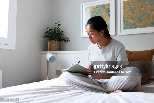 pensive young asian woman sitting on bed writing on journal in cozy bedroom. copy space. - tagebuch stock-fotos und bilder