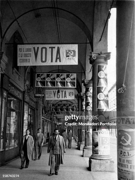 Passers-by walking under the porches covered with posters of the Italian Communist Party . Bologna, 1953