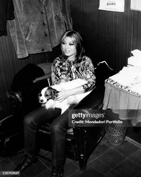 Smiling, Italian singer and actress Alida Chelli, born Alida Rustichelli, sits in an armchair and holds a little dog in her arms. Turin , 1968.