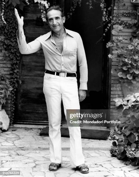 Actor Arnoldo Foà outside the entrance of his house smiling and waving, wearing summer clothes and open shoes. Rome, Italy, 1975.
