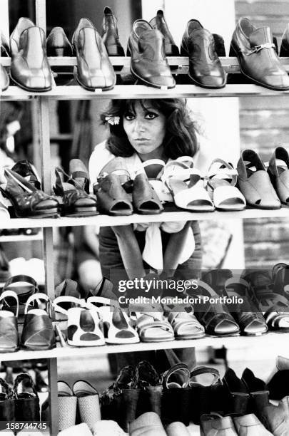 Ombretta Colli with a flower in her hair looking through some shelves full of shoes and sandals in a shoe shop. Pesaro, 1974.