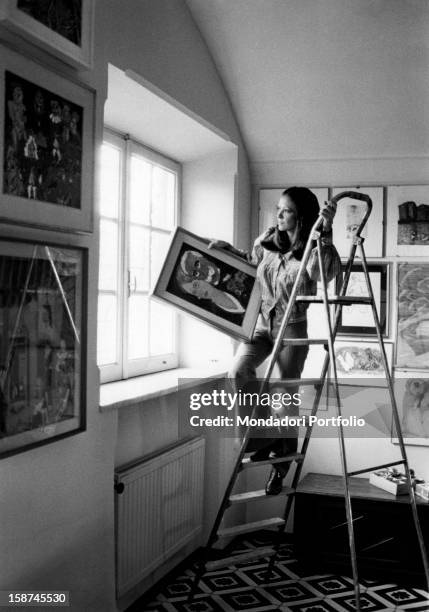 Italian actress and TV presenter Laura Efrikian going up a ladder and hanging a painting. Rome, 1970s