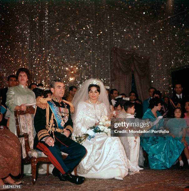 Mohammad Reza Pahlavi, Shah of Iran, is seated next to her third wife Farah Diba on the day of their marriage; her gown is by Yves St Laurent and on...