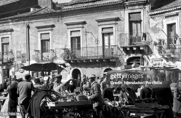 Sicilian men and women crowding the stalls of a street market. Sicily, March 1958