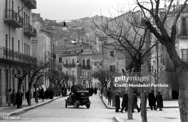 Many Sicilian people walking on the pavements of a tree-lined road. Sicily, March 1958