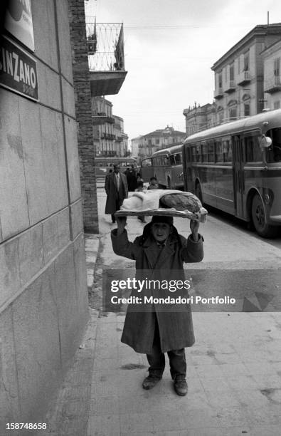 Sicilian kid walking carrying something on his head. Sicily, March 1958