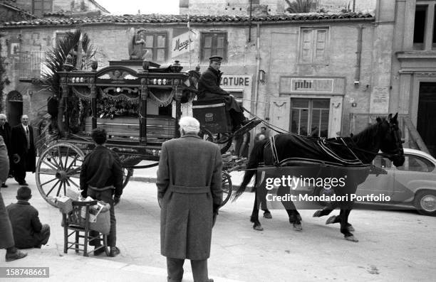 Some Sicilian people watching an ancient horse-drawn hearse. Sicily, March 1958