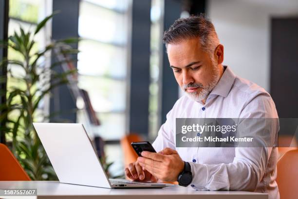portrait of a mature businessman with laptop looking at his mobile phone - digitale authenticatie stockfoto's en -beelden