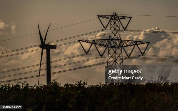 Wind turbine generates electricity besides electrical pylons carrying electricity cables across fields on July 16, 2023 near Bath, England. The UK...