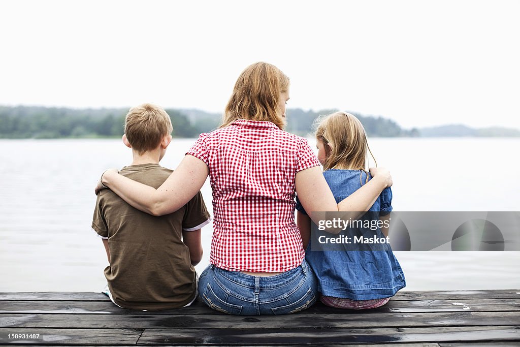 Rear view of mother with arm around her children sitting on pier