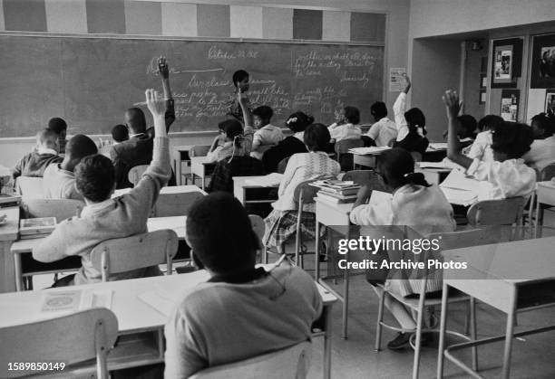 View from the back of a classroom, with children sitting at desks, some with their arms raised as a teacher stands at the head of the class before...