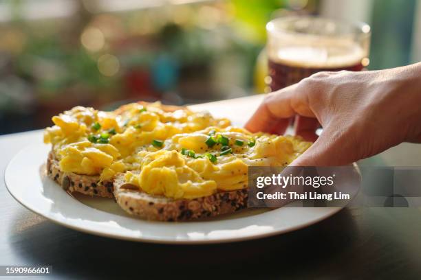 a cropped image of a woman's hand holding a piece of toasted bread with scrambled eggs on top, as she enjoys a healthy breakfast in the morning - egg white stock pictures, royalty-free photos & images