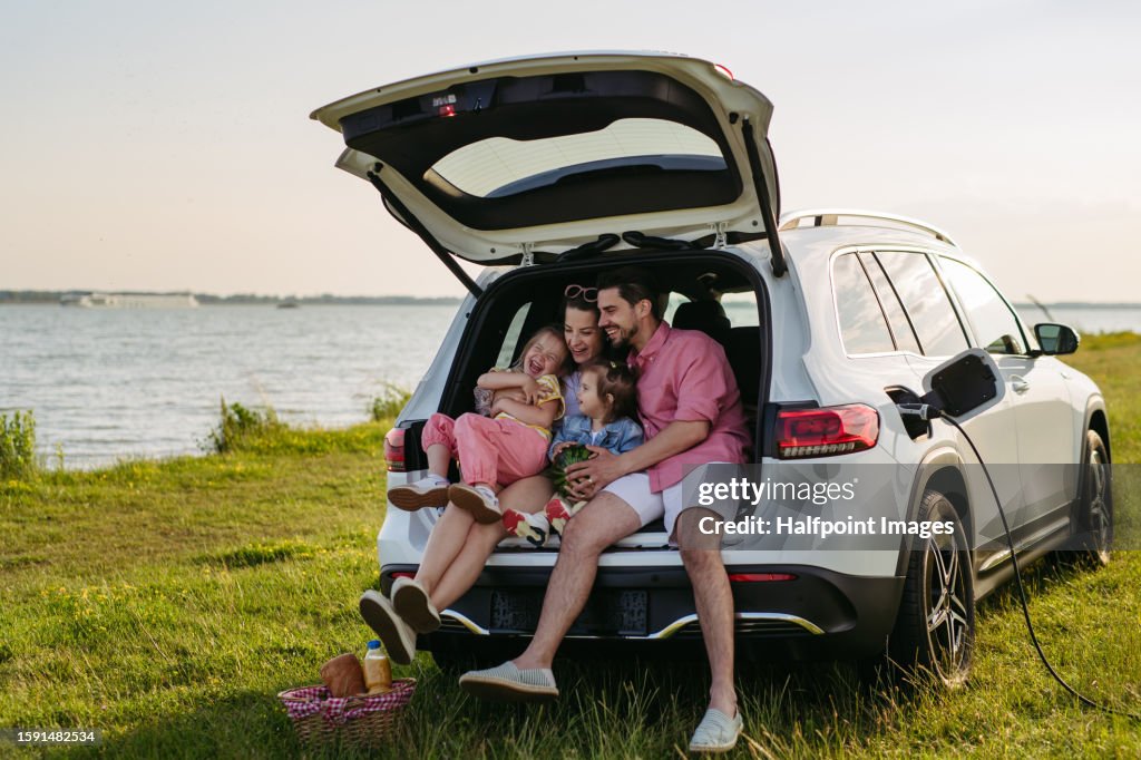 Happy family on the summer beach holiday sitting in the car trunk, waiting for electric car to charge.