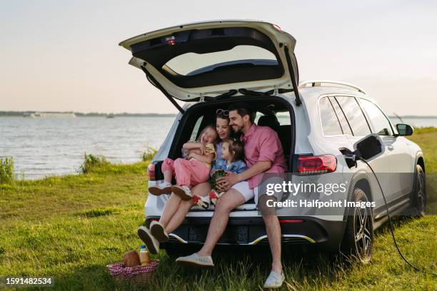 happy family on the summer beach holiday sitting in the car trunk, waiting for electric car to charge. - elektrische auto stockfoto's en -beelden
