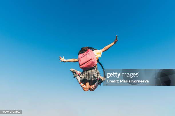 a carefree school girl with a pink backpack with outstretched arms jumps in the air against the blue sky. - back to school stock pictures, royalty-free photos & images