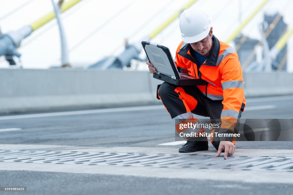 Using BIM Technology in the Construction Industry to Ensuring Compliance with Environmental Requirements. A male civil engineer in field operation holding a laptop to inspect the drainage and speed up the water flow of a water drain in a suspension bridge