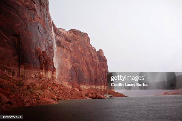rainstorm creating a waterfall flowing down  the red rock formation with houseboat at lake powell. - lake powell - fotografias e filmes do acervo