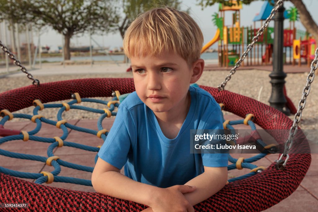 Loneliness on playground. Gloomy preschool blond boy (5-6 years) on playground swing looking away.