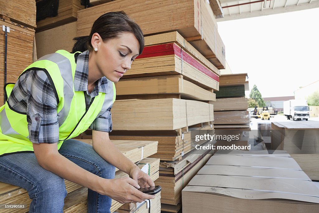 Female industrial worker using cell phone while sitting on stack of wooden planks