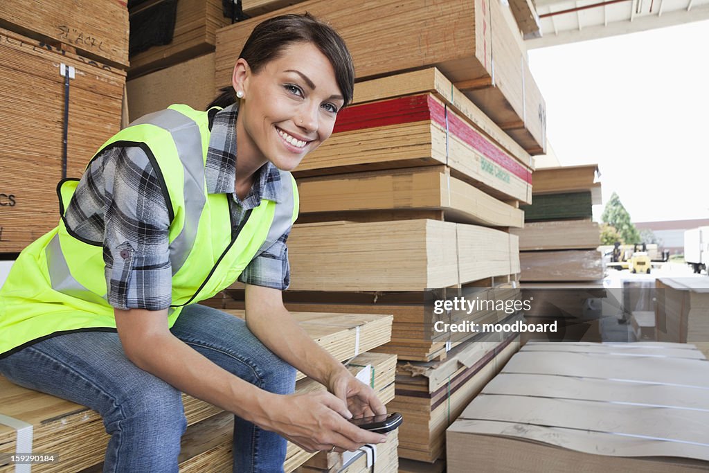 Portrait of female industrial worker using cell phone while sitting on stack of wooden planks