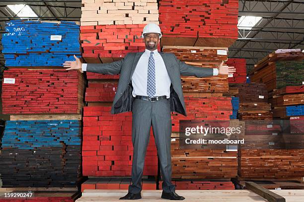 portrait of happy african american male engineer standing with arms outstretched in front of stacked wooden planks - erfolgreicher holzhandel stock-fotos und bilder