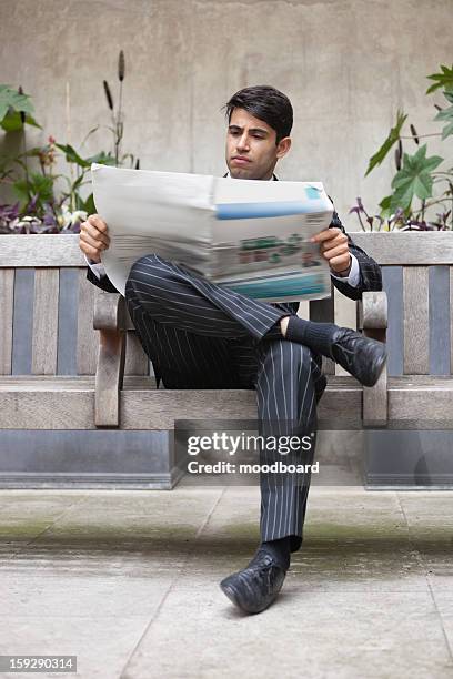 young indian businessman reading newspaper while sitting on bench - striped suit foto e immagini stock