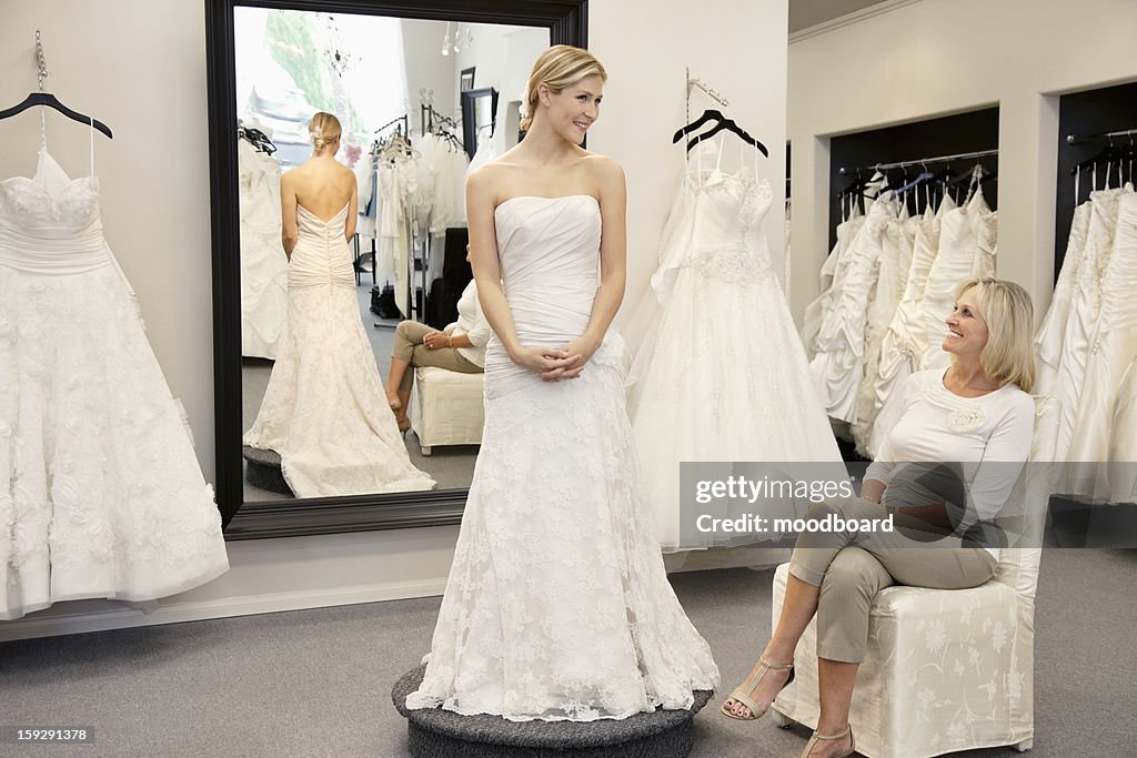 Happy mother looking at young daughter dressed in wedding gown in bridal boutique