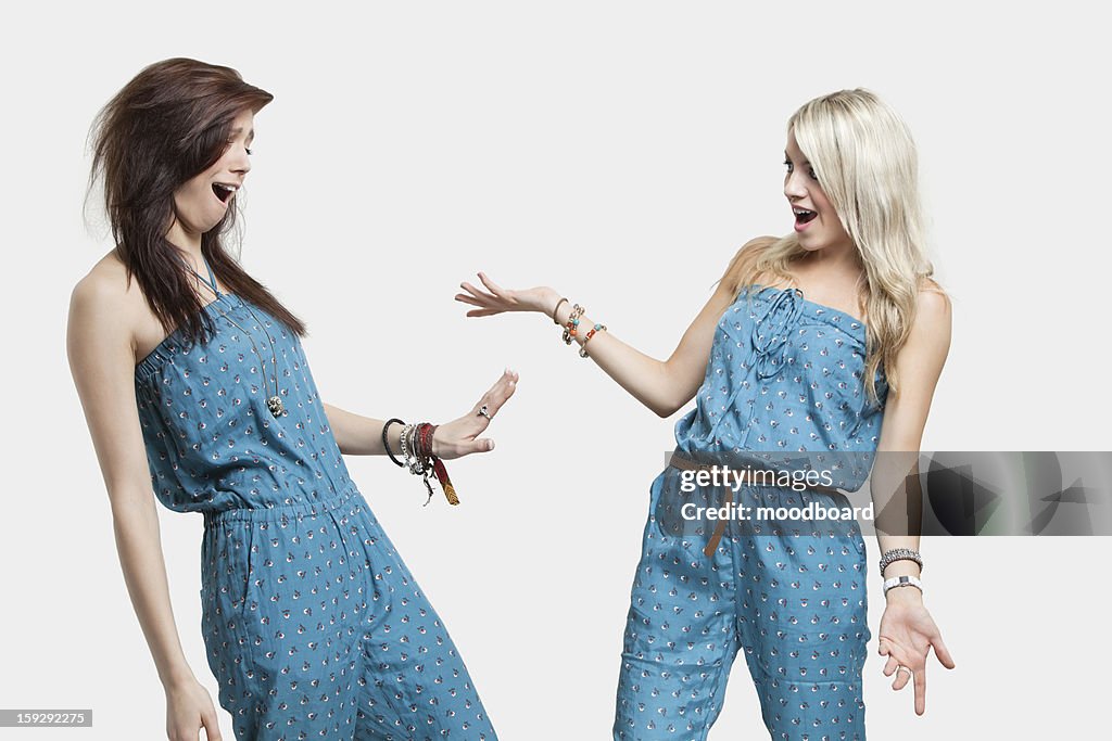 Two surprised women wearing similar jump suits looking at each other over gray background