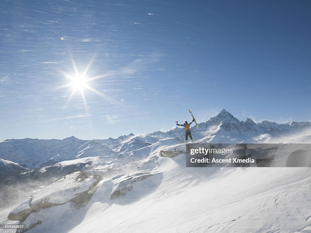 Skier stands on summit as wind blows snow,arms out