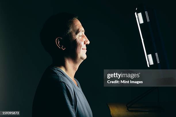 smiling middle aged man sitting in front of a light therapy box, a full spectrum light box which mimics the sun, and treats people suffering from seasonal affective disorder. - caixa de luz imagens e fotografias de stock